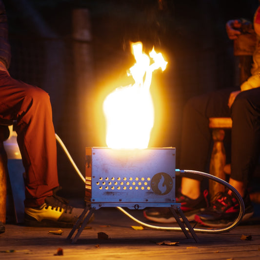 Two people sitting around a small stove with flames, likely in a camping or outdoor setting.